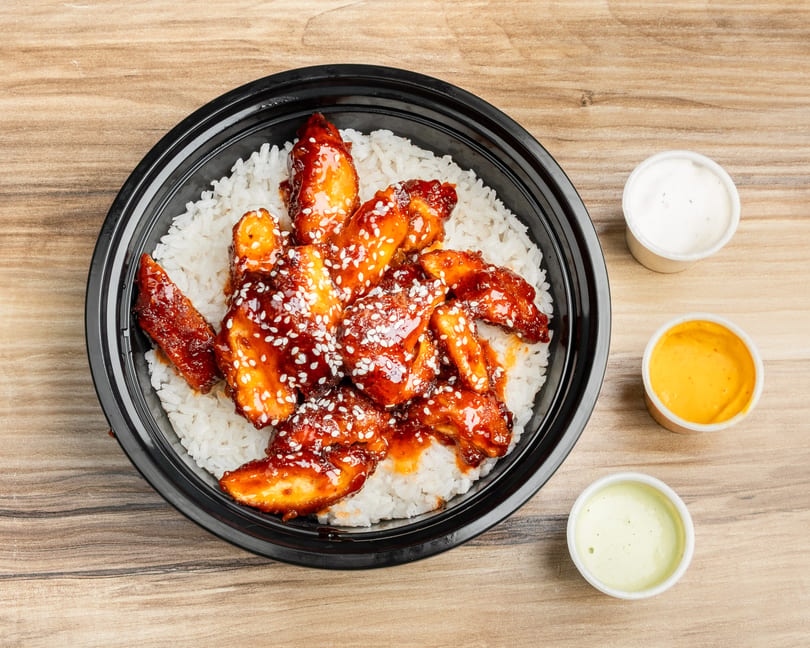 Sweet and sour chicken with sesame seeds over white rice in a black bowl, with three dipping sauces.