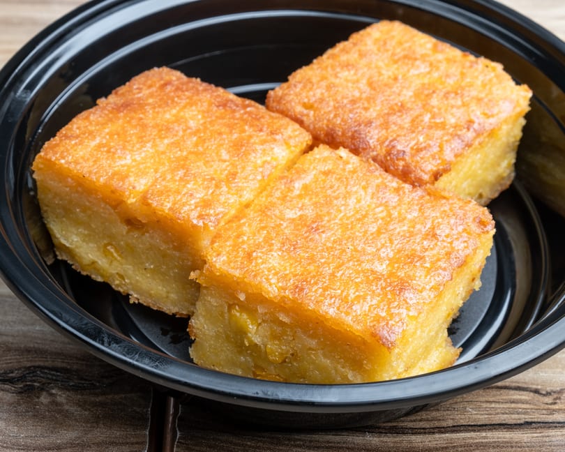 Three golden-brown squares of baked cassava cake with a slightly crispy top, in a black bowl.